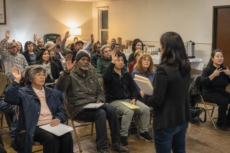 Residents seated in folding chairs at a community meeting, several raising their hands.